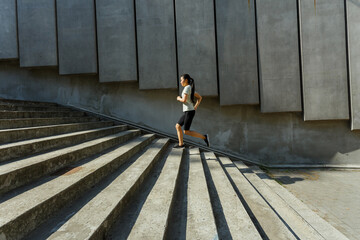 Long haired Asian woman in tracksuit runs up stone stairs past wall with concrete panels training on city street side view - 463840425