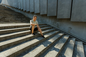Obraz na płótnie Canvas Attractive Asian woman in tracksuit and sneakers sits resting on empty grey steps casting shadow after outdoor training in sunny city