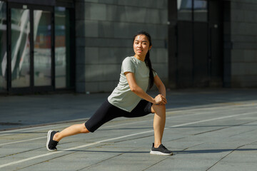 Fototapeta premium Positive strong Asian woman in tracksuit and shoes does forward dynamic lunges training on sunny city square on summer day