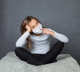 Portrait of a young teen girl in a medical mask with finger on her nose sitting on the ground with grey background. - 463842802