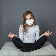 Portrait of a young teen girl in a medical mask sitting on the pillow in meditative position with grey background. - 463842804