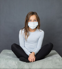 Portrait of a young teen girl in a medical mask sitting with crossed legs on the ground with grey background. - 463842805