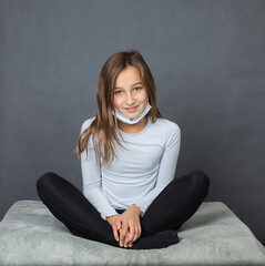 Portrait of a young teen girl smiling in a medical mask sitting on the ground with grey background. - 463842806