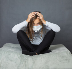 Portrait of a young teen girl in a medical mask with hands on her head sitting on the ground with grey background. - 463842812