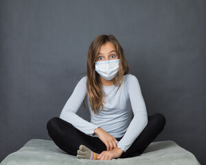 Portrait of a young teen surprised girl in a medical mask sitting on the ground with grey background. - 463842813
