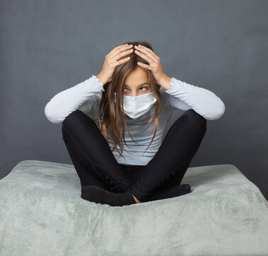 Portrait of a young teen girl in a medical mask with hands on her head sitting on the ground with grey background.