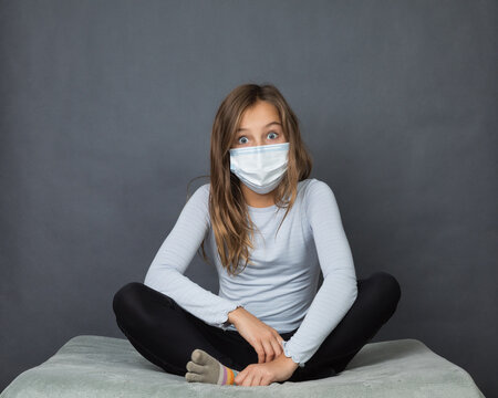 Portrait of a young teen surprised girl in a medical mask sitting on the ground with grey background.
