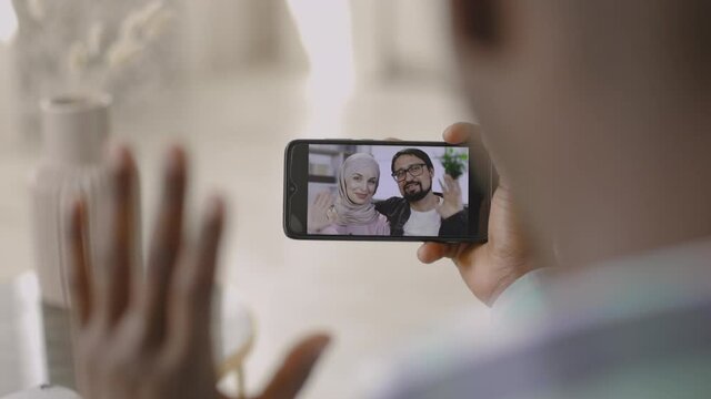Webcam conference, technology concept. Young African man at home, waving and talking on video call on phone with friends, joyful millennial Muslim Arab couple. Over the shoulder view of phone screen