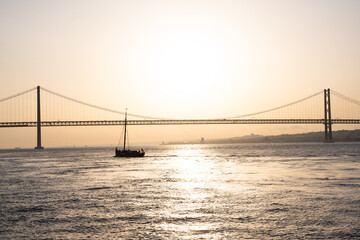 Fototapeta na wymiar Sailing boat floating next to a bridge during sunset in Lisbon