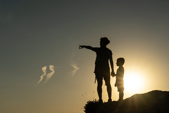 Mother and son against the light silhouetted by the sun pointing with their arms towards the horizon at sunset