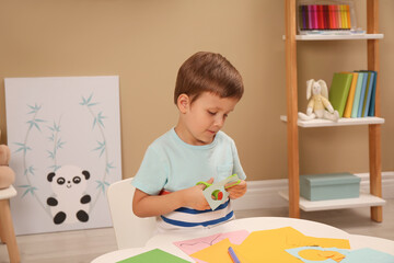 Little boy cutting color paper with scissors at table indoors