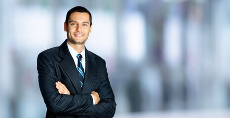 Portrait image of smiling brunette businessman in black suit, white shirt and blue stripes tie, standing in crossed arms pose, against blurred modern office interior background. Confident business man