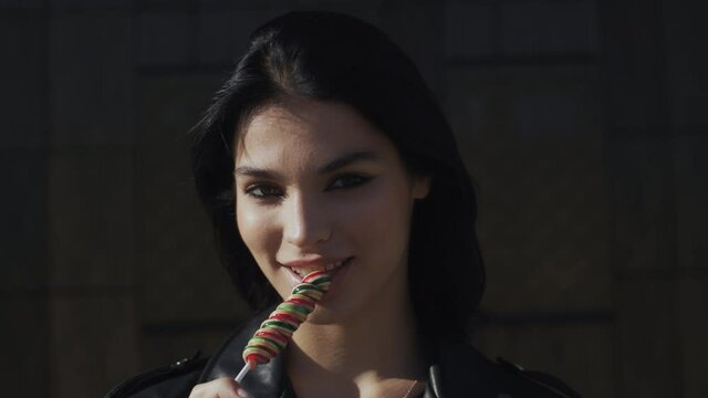 Portrait happy young brunette woman in trendy stylish look wearing black leather jacket seductively licks colorful lollipop candy looking at camera and smiling standing by wall in city street