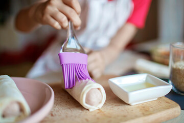 Fototapeta na wymiar Cute girl brushing sheet of filo pastry with melted butter, she cooks roasted sausage rolled in phyllo dough, tasty snack for kids, little chef enjoys cooking at home