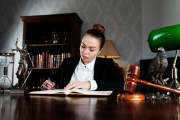 Young female lawyer during work in chamber. Gavel and Themis statue  on the brown shining desk.