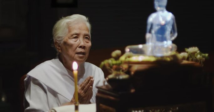 Asian Buddhist old woman with white hair white dress praying and raise their hands in front of the glass Buddha image and light candles in the night.	