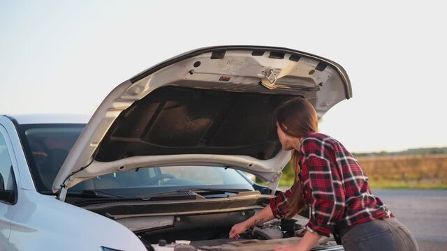 Car breakdown on road. Young puzzled woman looks under hood of car. Rear view.