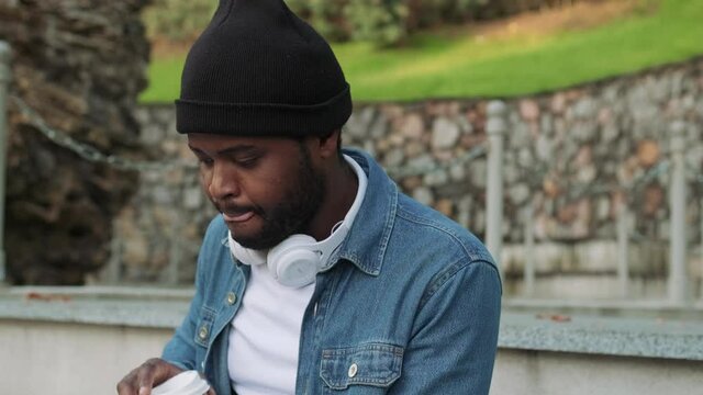 Concentrated African man in headphones drinking coffee on a concrete bench