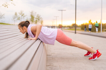 Side view of young sportive woman wearing activewear training doing push ups using street bench in...