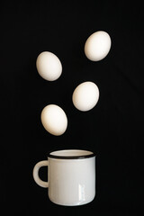 four white chicken eggs and a white mug on a black background. modern layout. minimalism. levitation white eggs fly over a white mug. modernism and symbolism