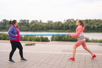 Fototapeta premium Wide shot of fat young woman doing squats exercises using resistance band for weight loss with personal trainer outdoor in cloudy summer evening. Instructor help overweight woman lose weight.