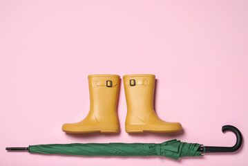 Pair of yellow rubber boots near green umbrella on pink background, flat lay. Space for text
