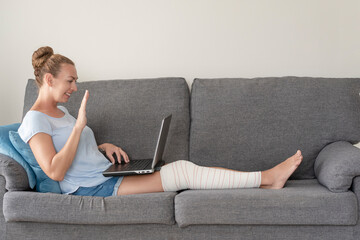 a young woman sitting on the sofa with leg in bandage , video conference concept