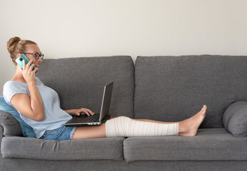 a young woman sitting on the sofa with her leg in bandage talking on the cell phone and using the...
