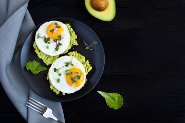 Top view of avocado toasts for breakfast with rye bread, avocado puree, fried eggs and microgreen on the black  background. Copy space.