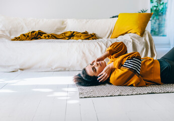 Woman in casual clothing with headphones lying down the floor in her living room and enjoys listening the music. Leisure time of single woman relaxing in isolation streaming songs from player. - obrazy, fototapety, plakaty