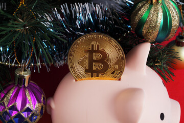 piggy bank with gold bitcoin under a christmas tree decorated with christmas balls and tinsel on a red background, close-up
