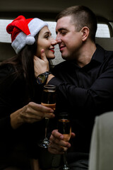 Happy young couple in a car. Man and woman celebrating Christmas with champagne in car. Woman wearing Santa's hat. Sensual lovers in black clothes.