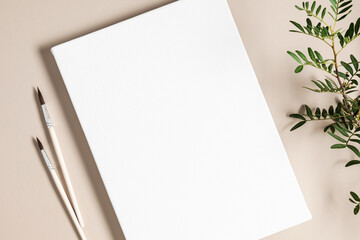Blank white canvas mockup with paint brushes and green leaves on beige background. Flat lay, top...