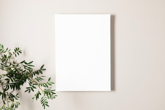 Blank white canvas mockup and green leaves on beige background. Flat lay, top view, copy space