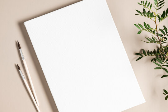 Blank white canvas mockup with paint brushes and green leaves on beige background. Flat lay, top view, copy space