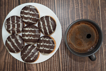 a black mug with natural coffee stands on a wooden table next to a white plate with chocolate cookies. top view