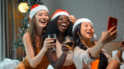 young girls European, Asian and African in red New Year's clothes and hats sit on a bed at a Christmas tree holding glasses of wine made photo on a phone, smiling and celebrating Christmas