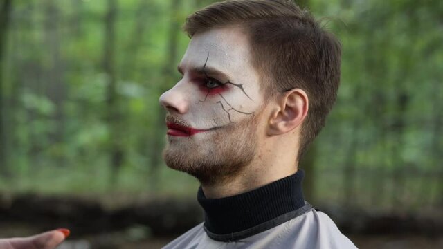 Close-up portrait of handsome young man in forest with unrecognizable visagiste making Halloween makeup on face. Caucasian guy getting ready for holiday celebration or performance