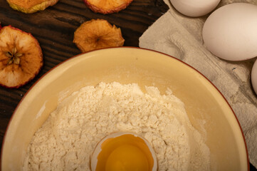 A broken white chicken egg in a bowl with wheat flour, dried apple chips and several whole eggs in a tray made of white cardboard, close-up selective focus.