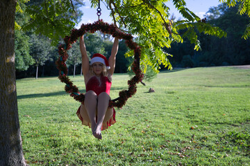 Young blonde woman in a red party dress and a Santa Claus hat sitting on an aerial hoop hanging from a tree. Christmas concept, Santa Claus, New Year's Eve party, aerial hoop, curvy girl.