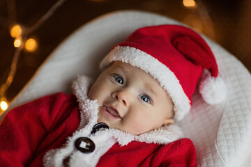 Closeup portrait of newborn baby. Cute Caucasian baby girl 4-5 months old in Santa costume lying on knitted cozy blanket in cocoon near decorated fir tree. Merry Christmas xmas and happy new year 2022