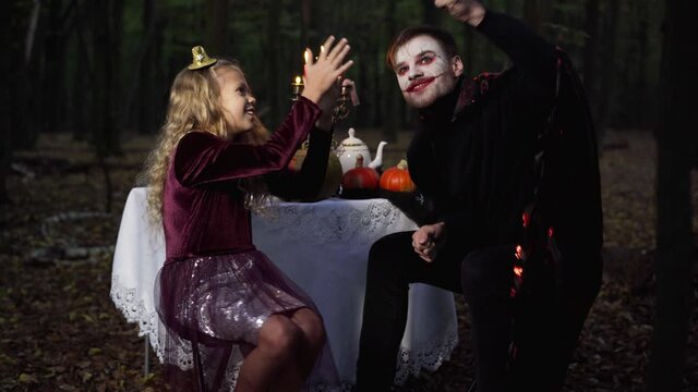Man with scary Halloween face paint holding lollipop playing with pretty girl in forest in darkness. Vampire and child on holiday in woodland outdoors having fun with sweet dessert