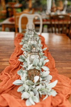 Terracotta color rag runner on the wooden table Wedding decorations Covered festive table Candle White flowers and green Glass vase Table set with candles and flowers for a wedding reception