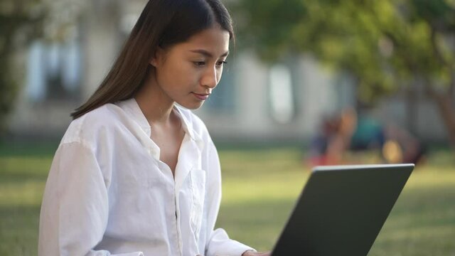 Close-up portrait of pretty asian college girl with laptop preparing for exam while sitting on campus lawn. Lovely brunette woman student in white blouse studying outside class in nature