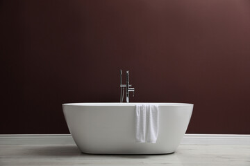 Modern ceramic bathtub with towel near burgundy wall indoors