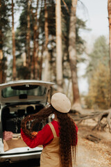girl with long hair and a white beret stands in the autumn forest against the backdrop of a large car