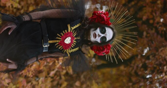 Vertical view of young woman posing with sugar skull makeup and red roses dressed in black costume of death as Santa Muerte against background of autumn forest. Day of the Dead or Halloween concept.