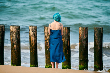 Young blue-haired woman in long dark blue dress standing on sandy beach looking at sea horizon