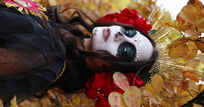 Vertical portrait of young woman with sugar skull makeup and red roses dressed in black costume of death as Santa Muerte in autumn forest. Day of the Dead or Halloween concept.