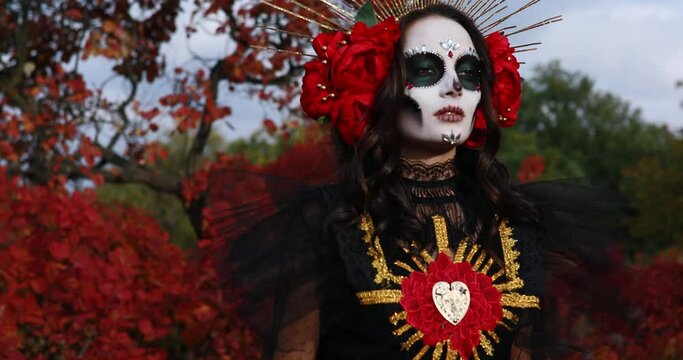 Young woman walks with sugar skull makeup and red roses dressed in black costume of death as Santa Muerte against background of autumn forest. Day of the Dead or Halloween concept.